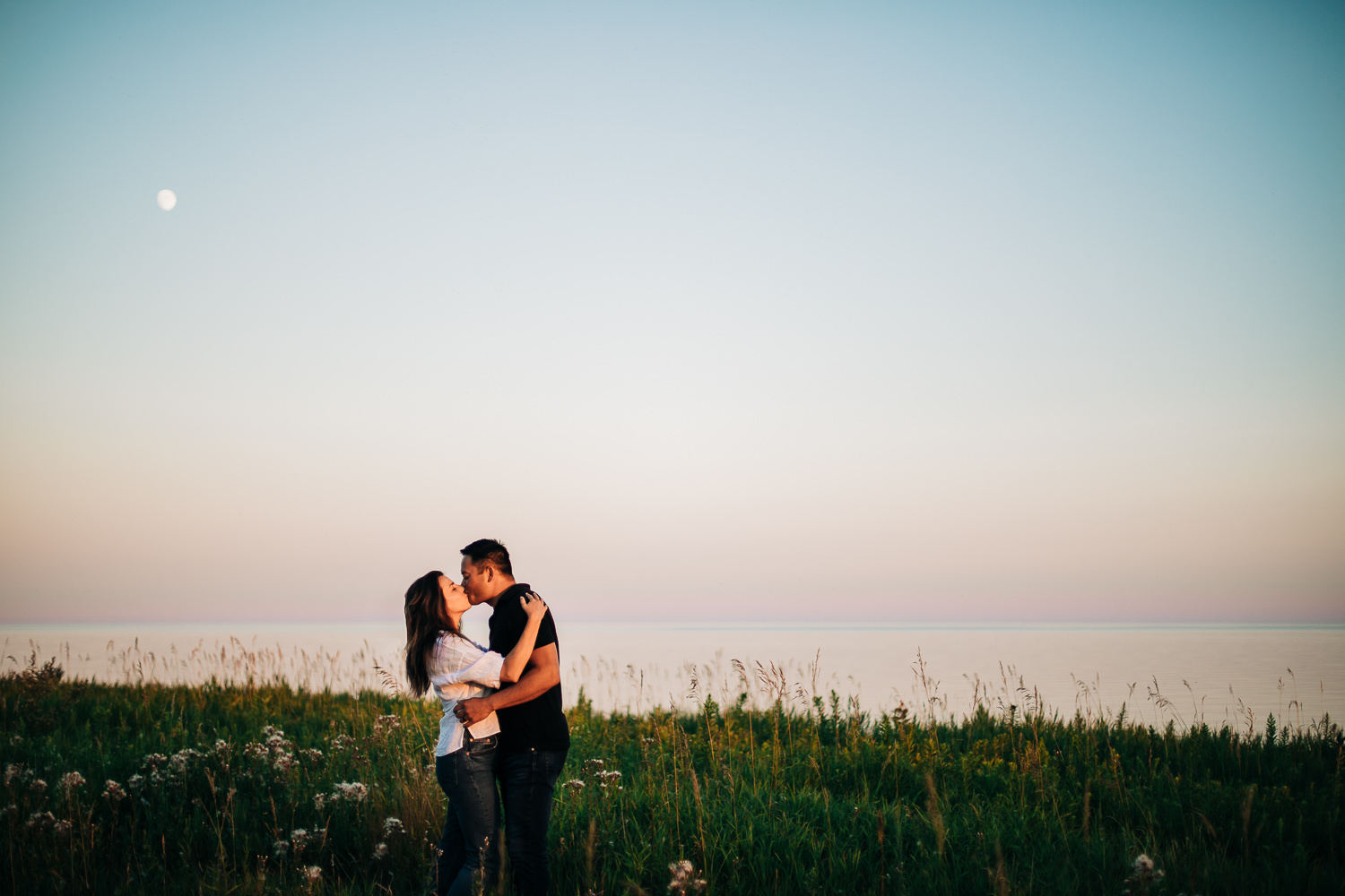 Couple embracing and kissing atop a cliff with wildflowers at sunset with visable moon