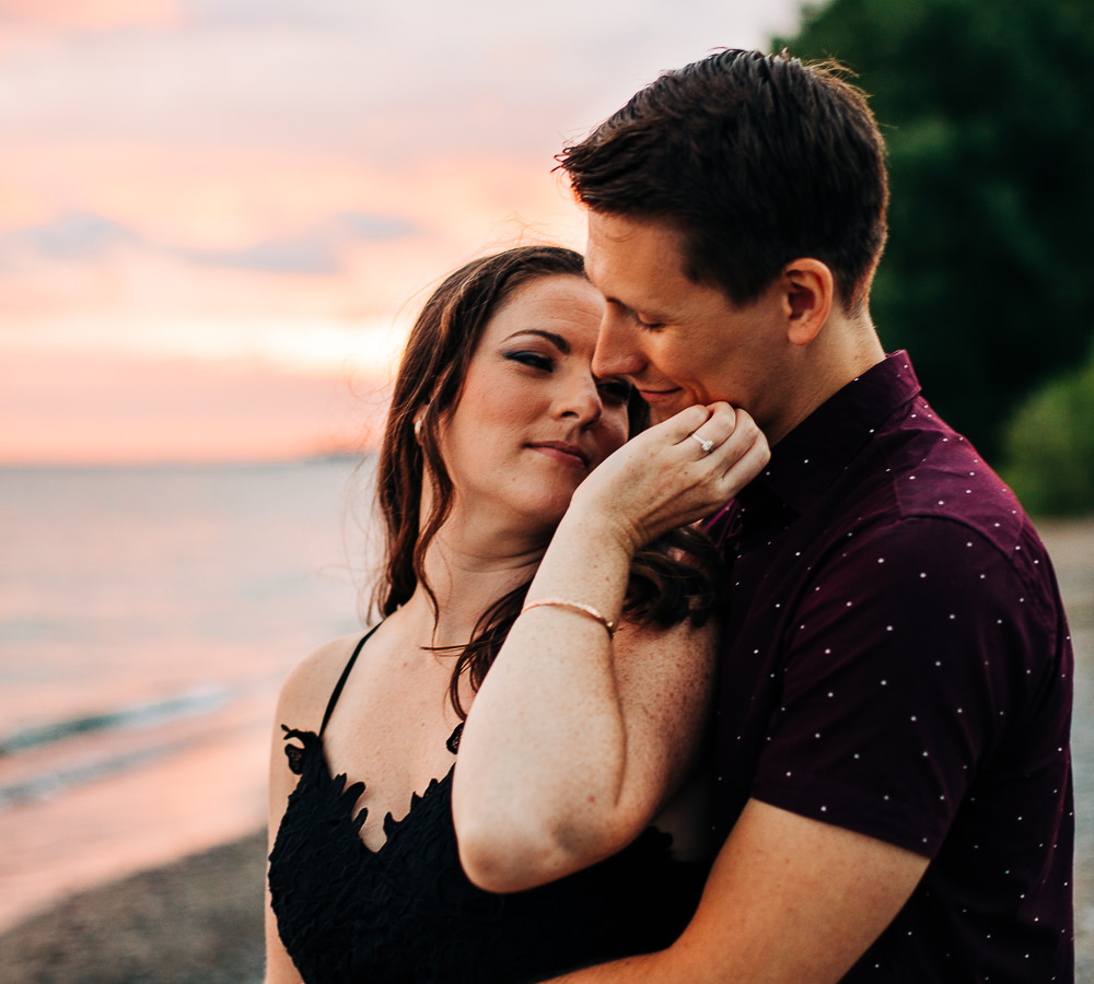 Man embraces his fiancé while she gently strokes his chin on a beach at sunset